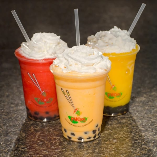 Three colorful Phở 95 bubble tea smoothies with whipped cream and straws on a dark surface.