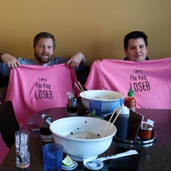 Two men at Phở 95 holding up pink "I am a Pho King LOSER" t-shirts above large pho bowls.