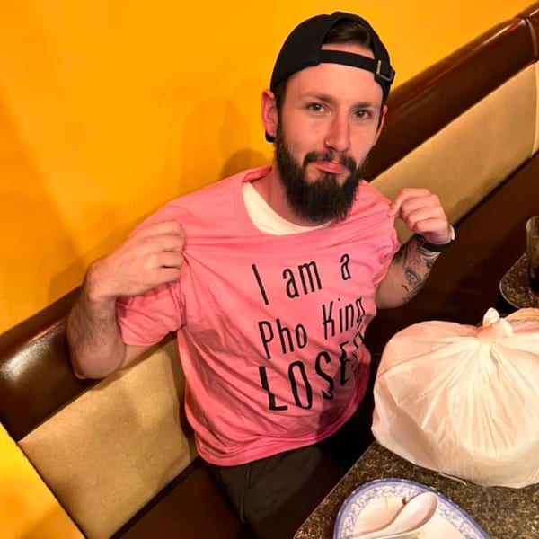 A man in a black cap and beard, wearing a pink "I am a Pho King LOSER" t-shirt, sits in a booth.