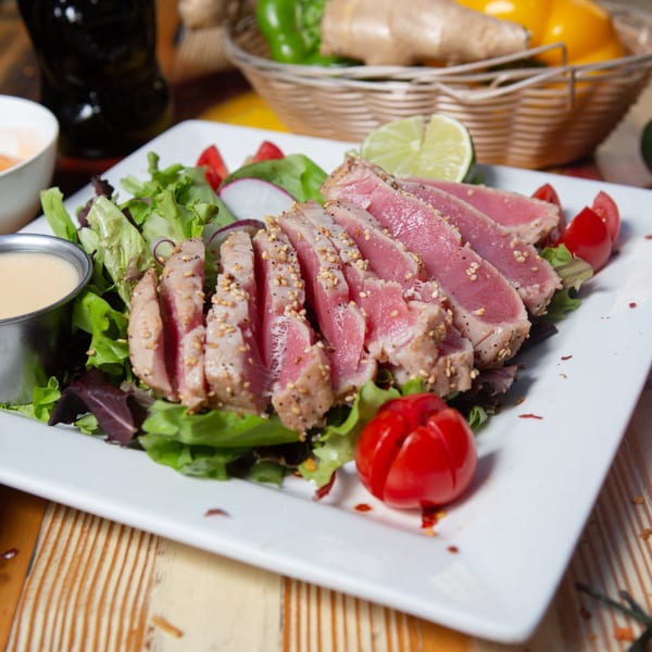 Seared tuna slices with sesame seeds on a bed of greens, cherry tomatoes, and a side of dressing.