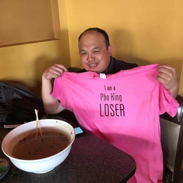 Man at Phở 95 holding up a pink "I am a Pho King LOSER" t-shirt next to a large bowl of pho.