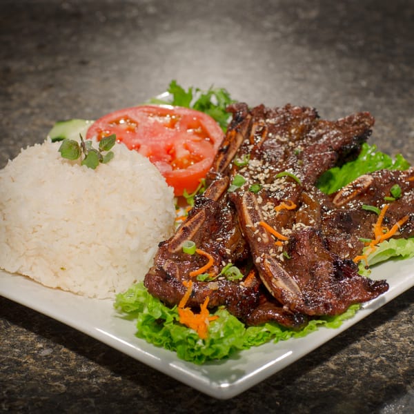 Grilled short ribs with rice, sliced tomatoes, and lettuce on a white rectangular plate.