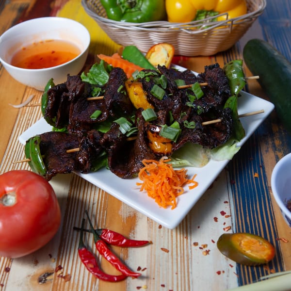 Grilled beef skewers with bell peppers, green onions, and shredded carrots, served with a dipping sauce.